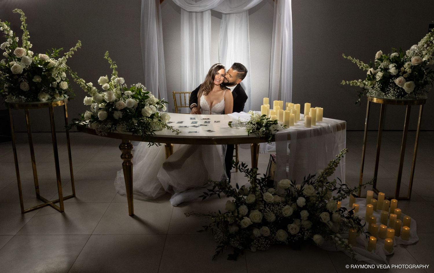 Bride and groom share a romantic moment at elegantly decorated sweetheart table with white floral arrangements and candles.