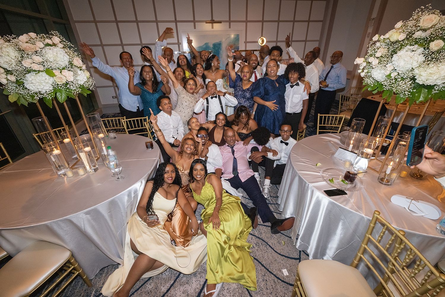 Large wedding reception group photo with guests cheering around elegantly decorated tables with tall floral centerpieces.