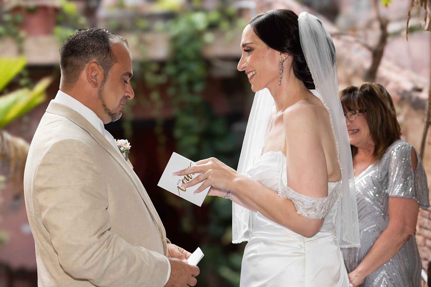 Bride and groom exchanging vows at outdoor wedding ceremony with officiant in silver dress in background.