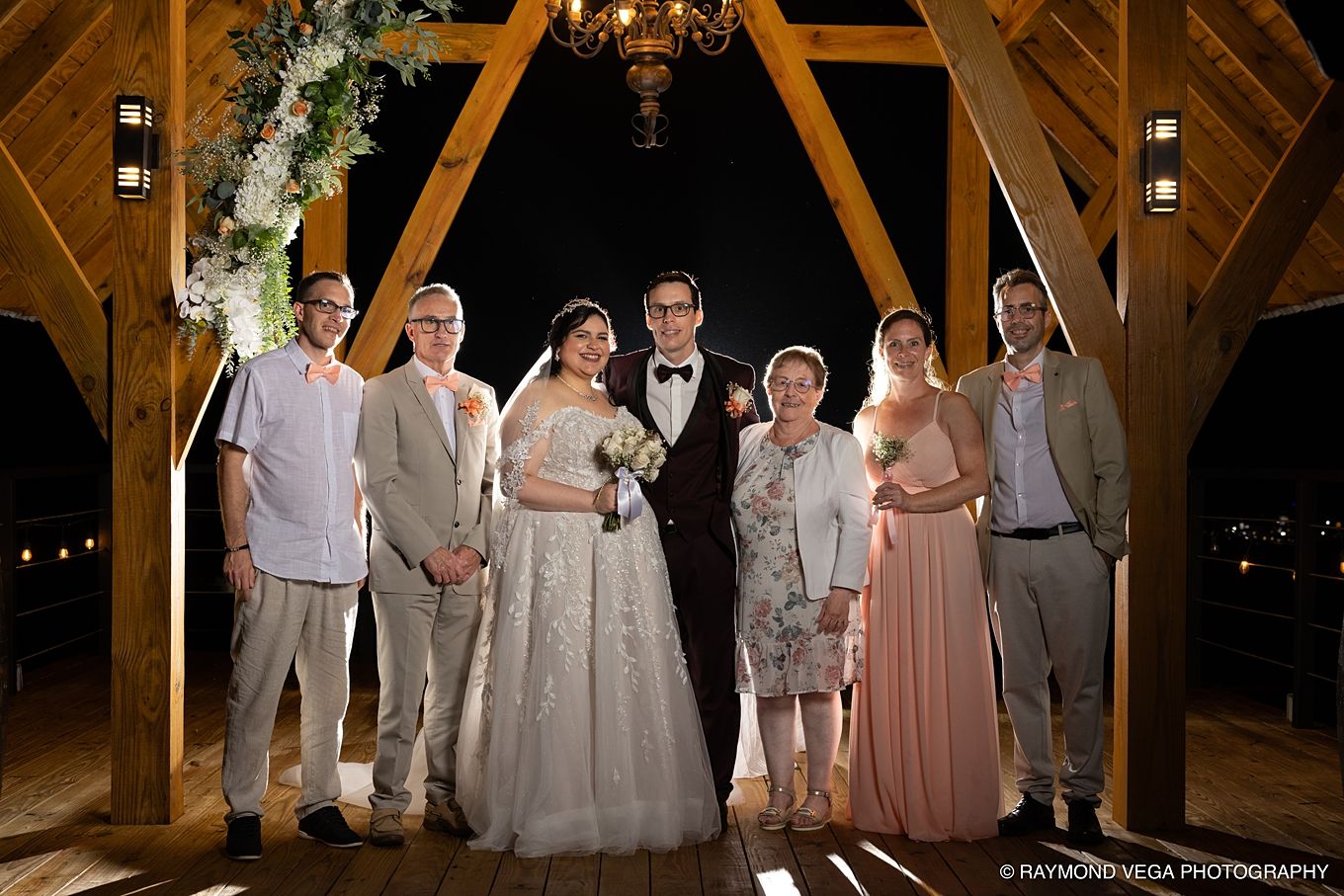 Wedding party poses under wooden arch at night, bride in white gown, groom in tuxedo, surrounded by family in formal attire.