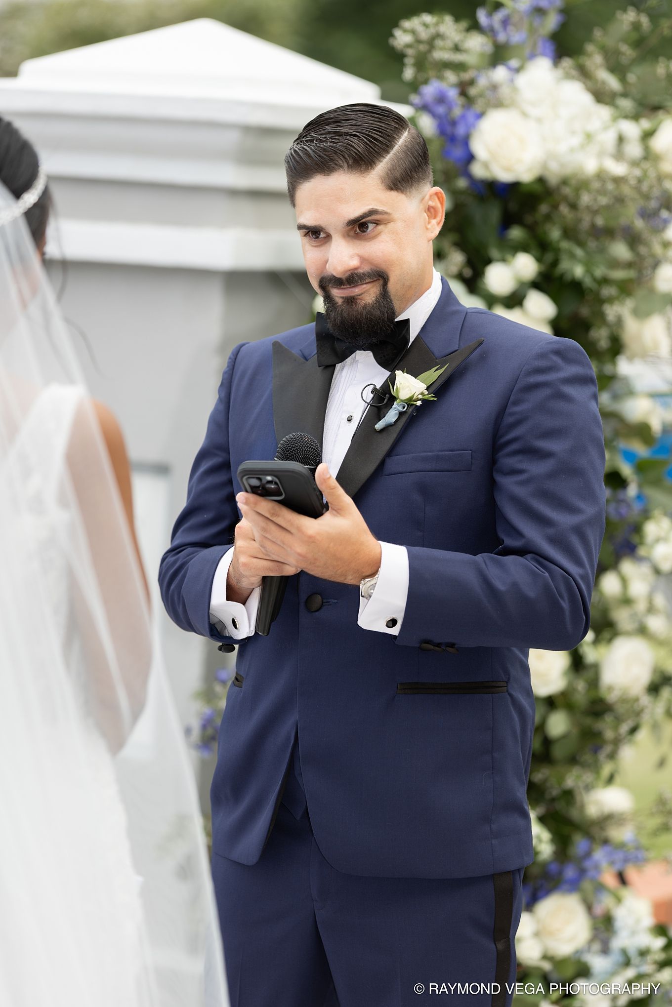 Groom in blue tuxedo smiling while holding vows and microphone at outdoor wedding ceremony with floral backdrop. Antiguo Casino Puerto Rico