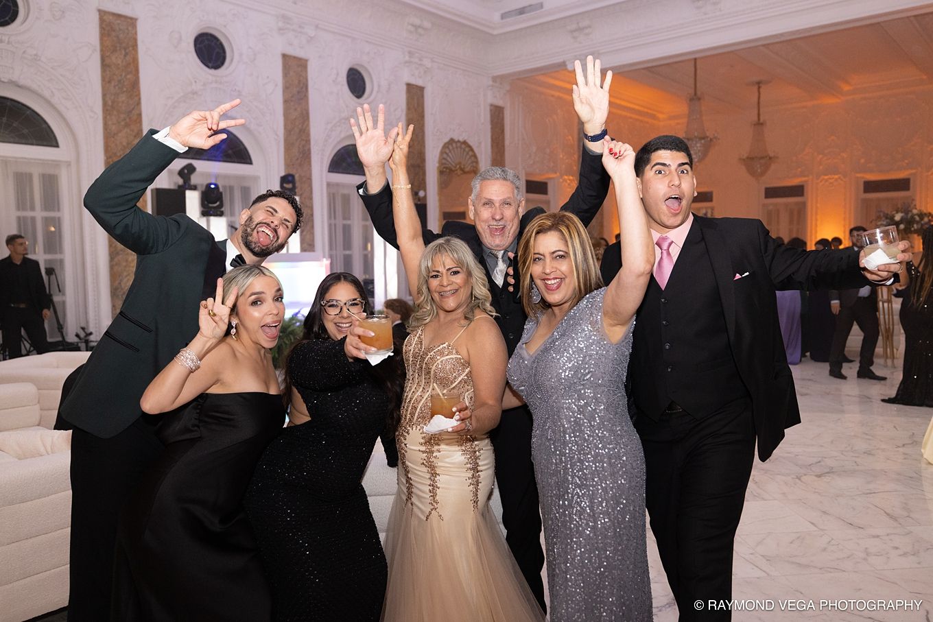 Group of elegantly dressed guests celebrating at a formal wedding reception, raising hands and smiling.