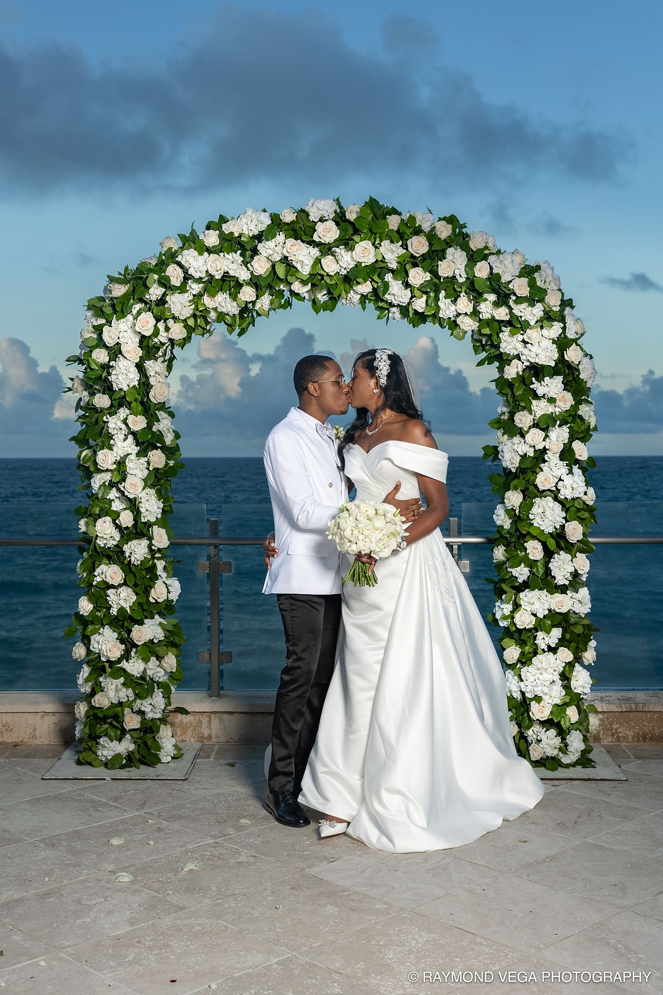 Newlywed couple kissing under a white floral arch by the ocean at their outdoor wedding ceremony.