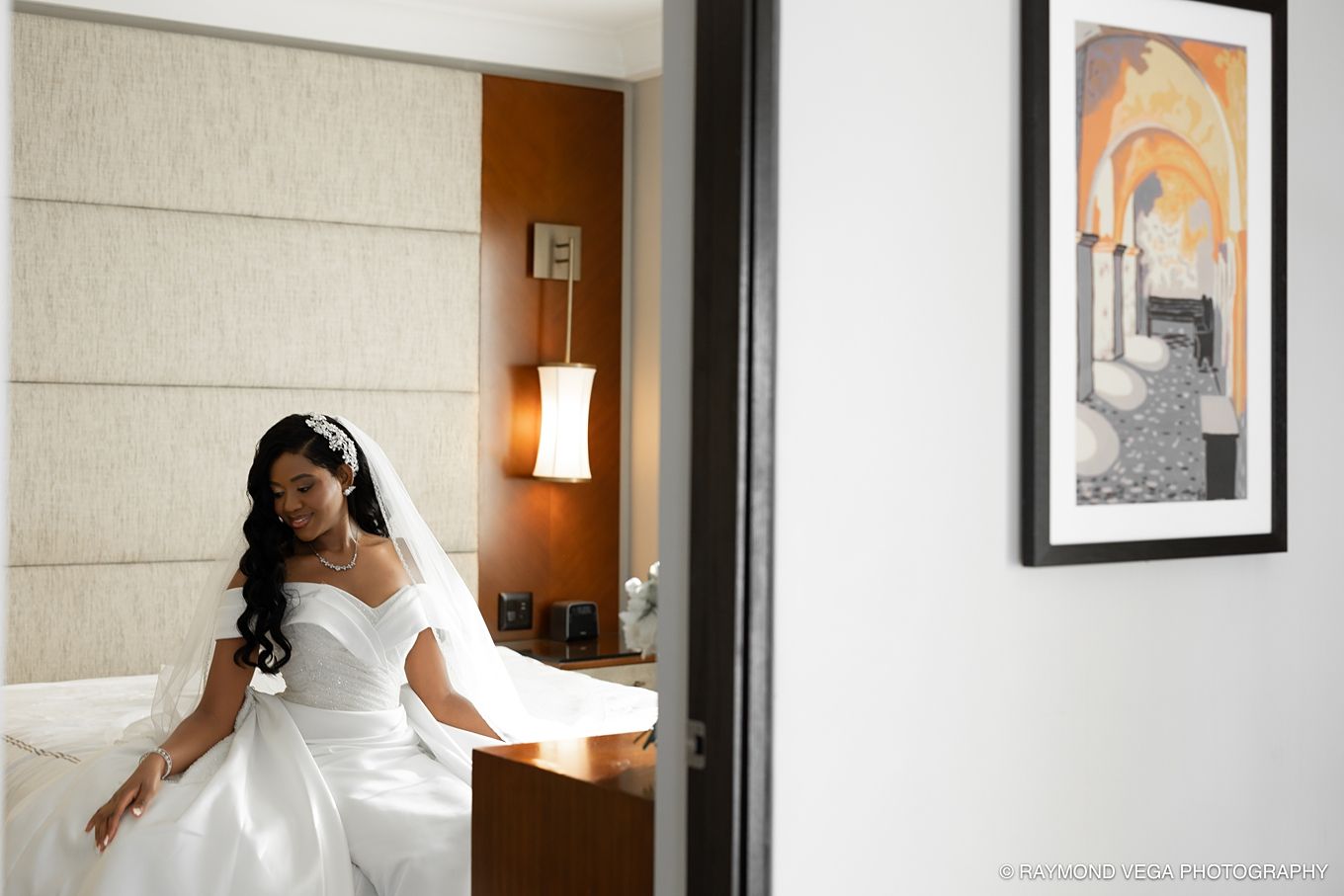 Bride in white wedding gown sitting on hotel bed, wearing headphones, with framed artwork visible in elegant room. Hotel Condado Vanderbilt