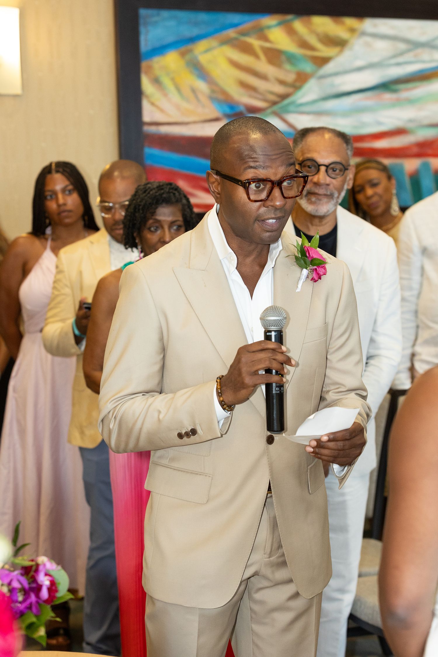 Man in tan suit speaking at wedding ceremony holding microphone and paper with guests in background.