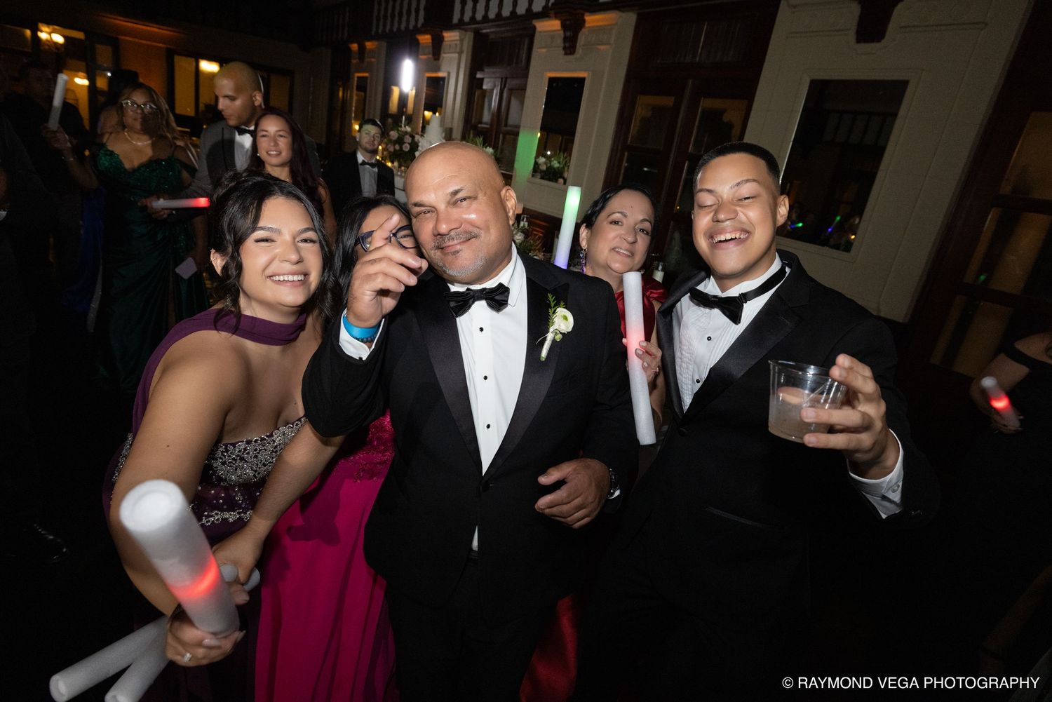 Guests in formal attire smile and raise drinks at a lively wedding reception party.