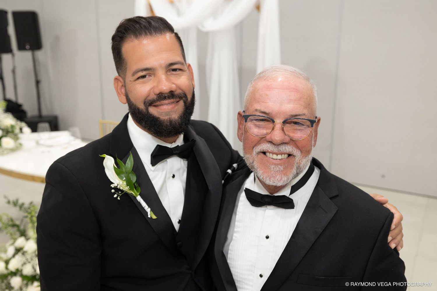Two men in tuxedos smiling together at a wedding ceremony, one wearing a boutonniere.