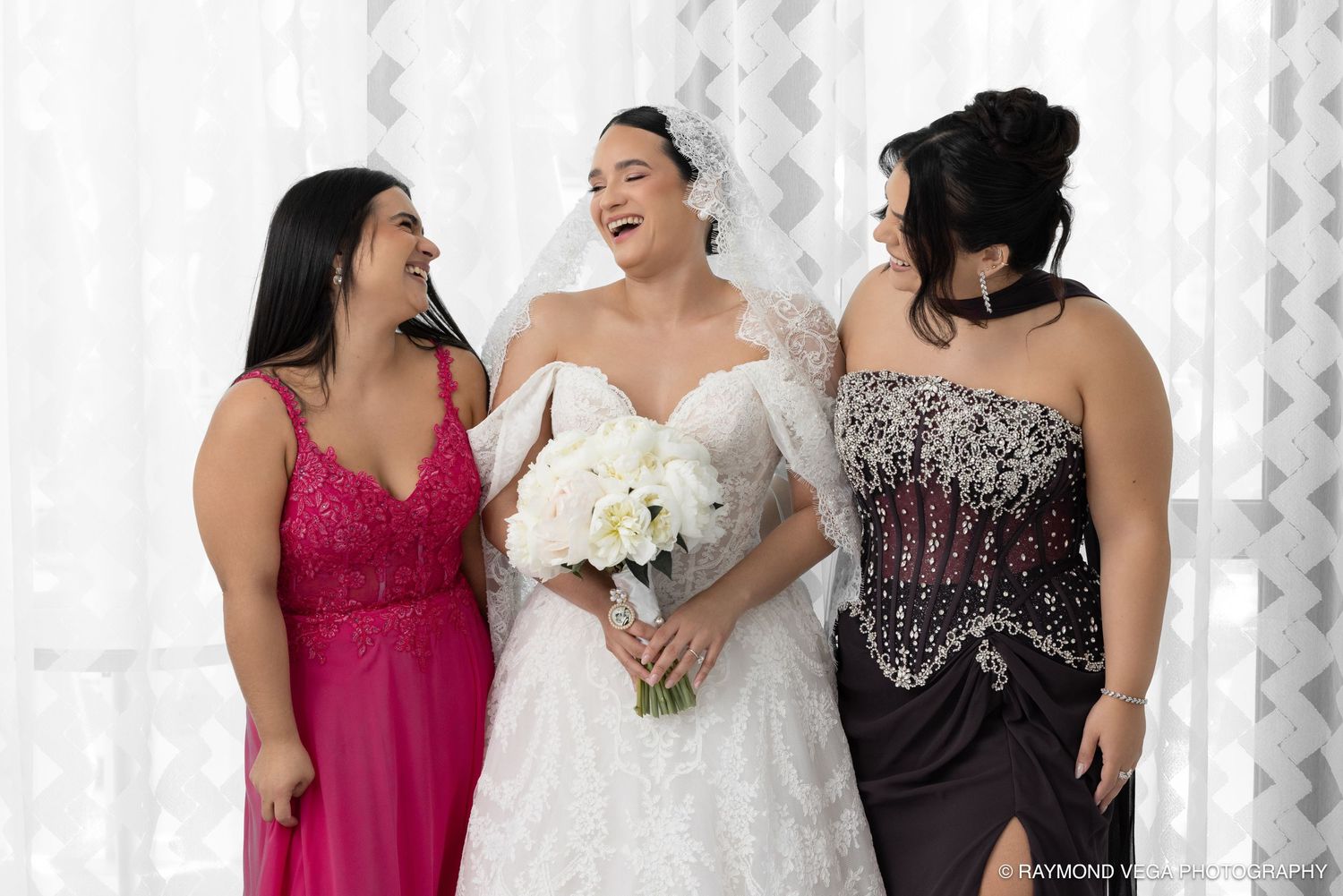 Bride in white gown holding bouquet laughs with two bridesmaids in pink and black dresses.