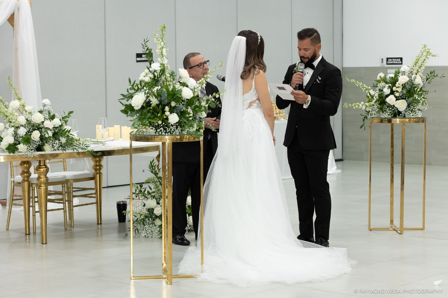 Bride and groom exchange vows at elegant wedding ceremony with white floral arrangements and gold stands.