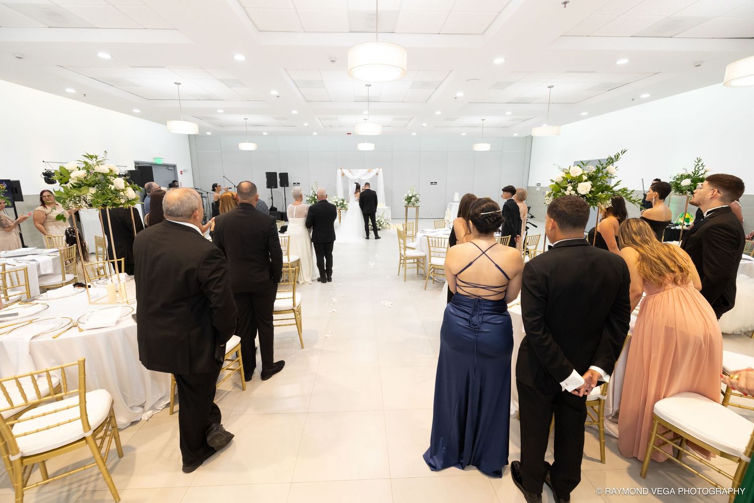 Elegant wedding reception hall with guests mingling, gold chairs, white floral arrangements, and a long aisle leading to the altar.