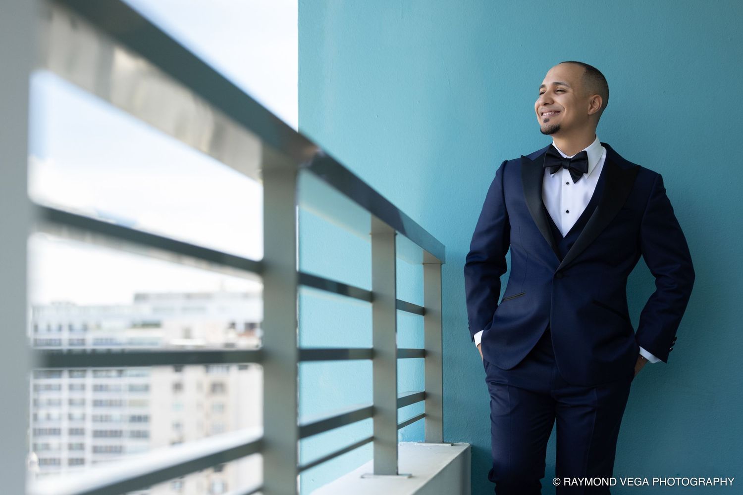 A man in a navy tuxedo with bow tie stands on a balcony, gazing confidently at a city skyline.