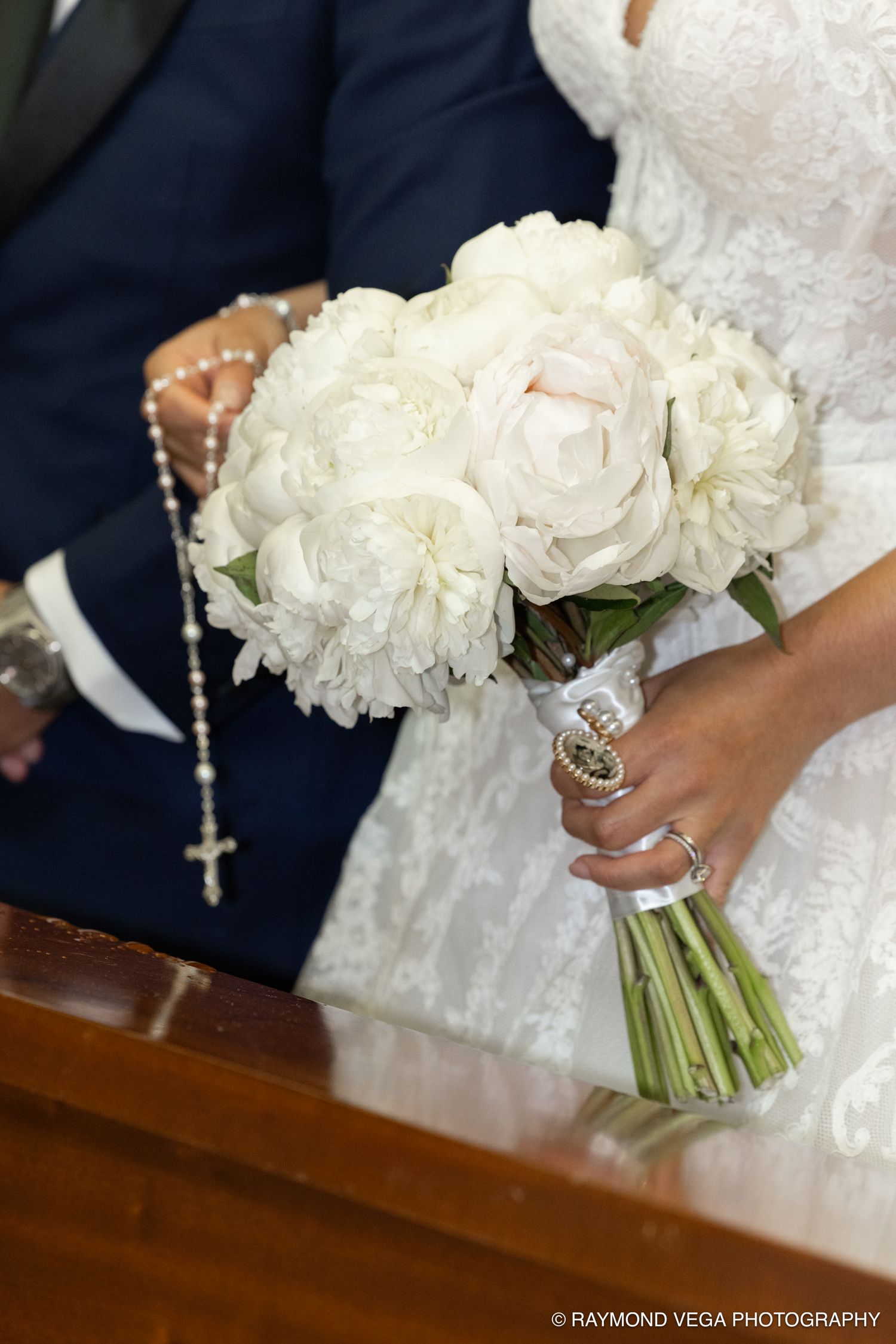 Bride holding a white peony wedding bouquet with a rosary during a church ceremony.