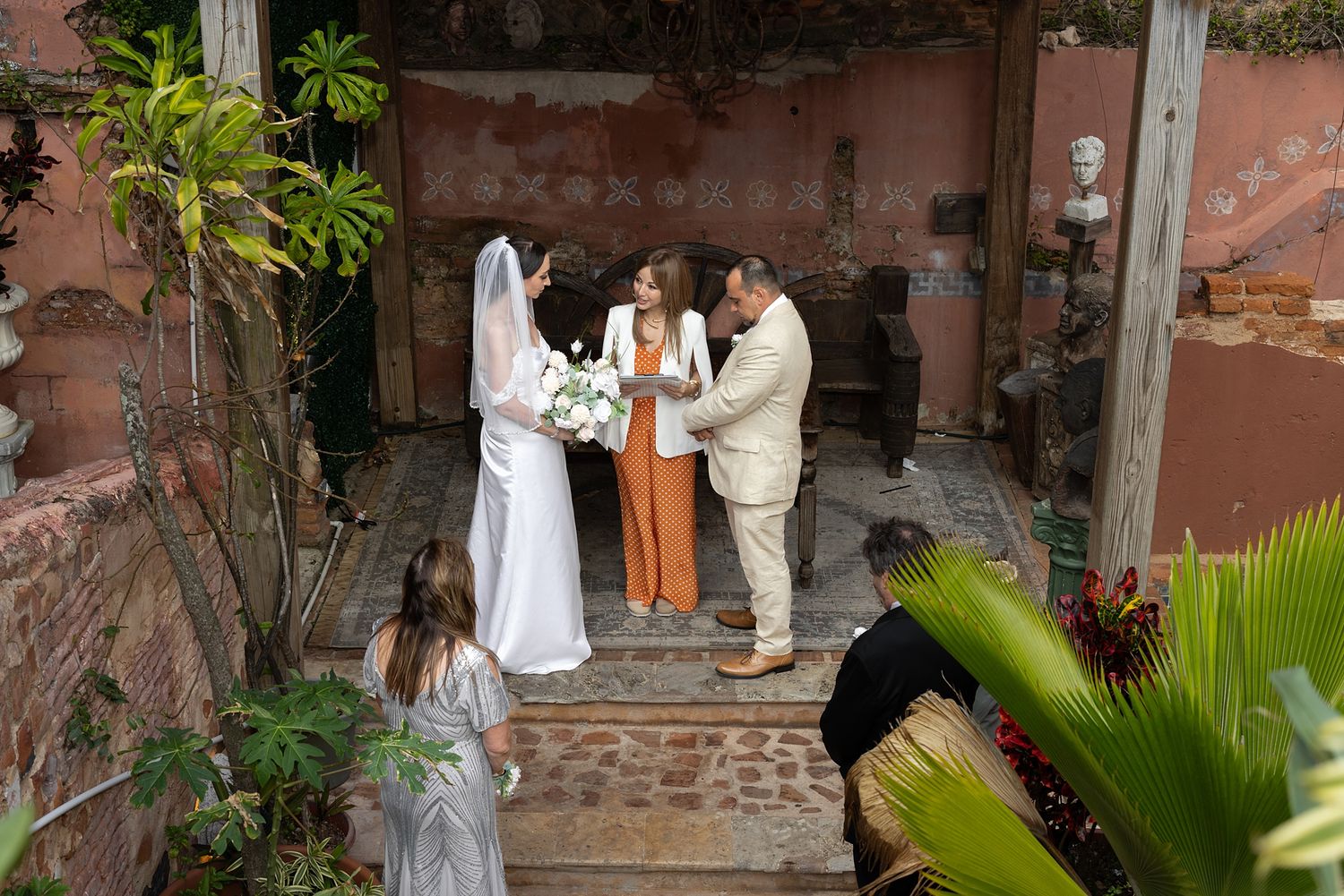 Outdoor wedding ceremony in a rustic courtyard with bride, groom, and officiant surrounded by lush greenery. AMR Destination Weddings