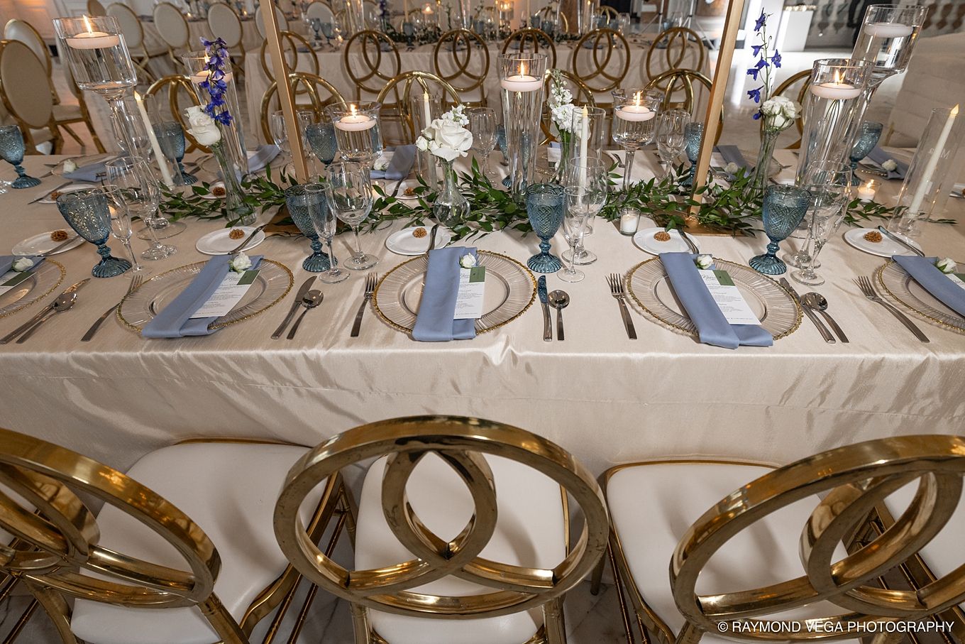 Elegant wedding reception table with gold ring chairs, blue napkins, candles, and floral centerpieces on white linen. Handcrafted Events