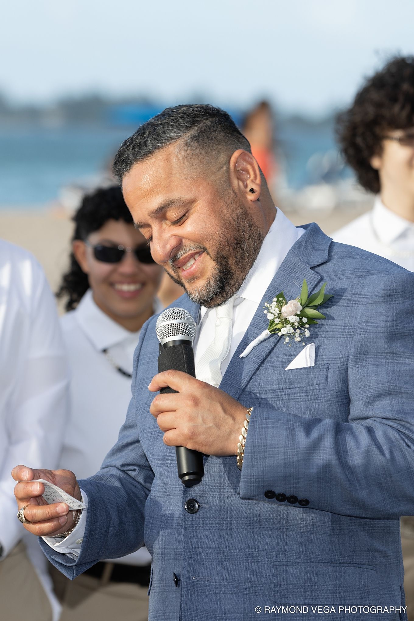 Man in blue suit speaking at beach wedding ceremony, holding microphone and notes, wearing boutonniere.