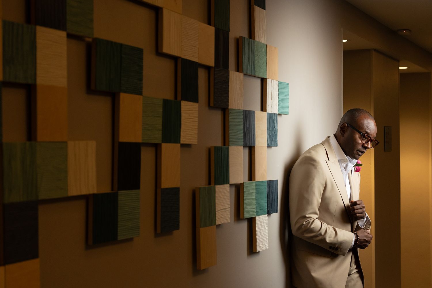 Elegant man in beige suit adjusting jacket, leaning against wall with colorful geometric art panels.