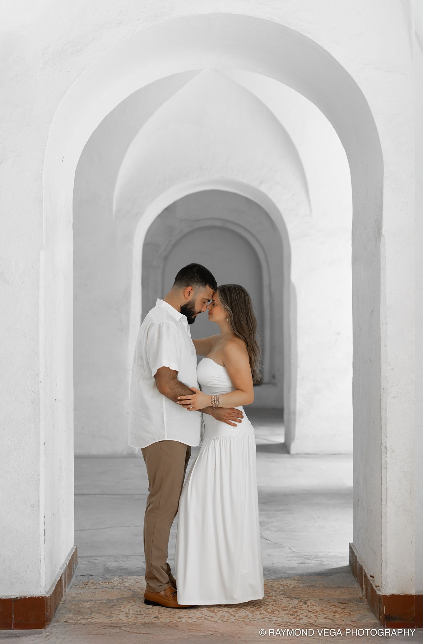Couple in white outfits sharing a romantic moment under elegant white arched hallway, captured by Raymond Vega Photography.