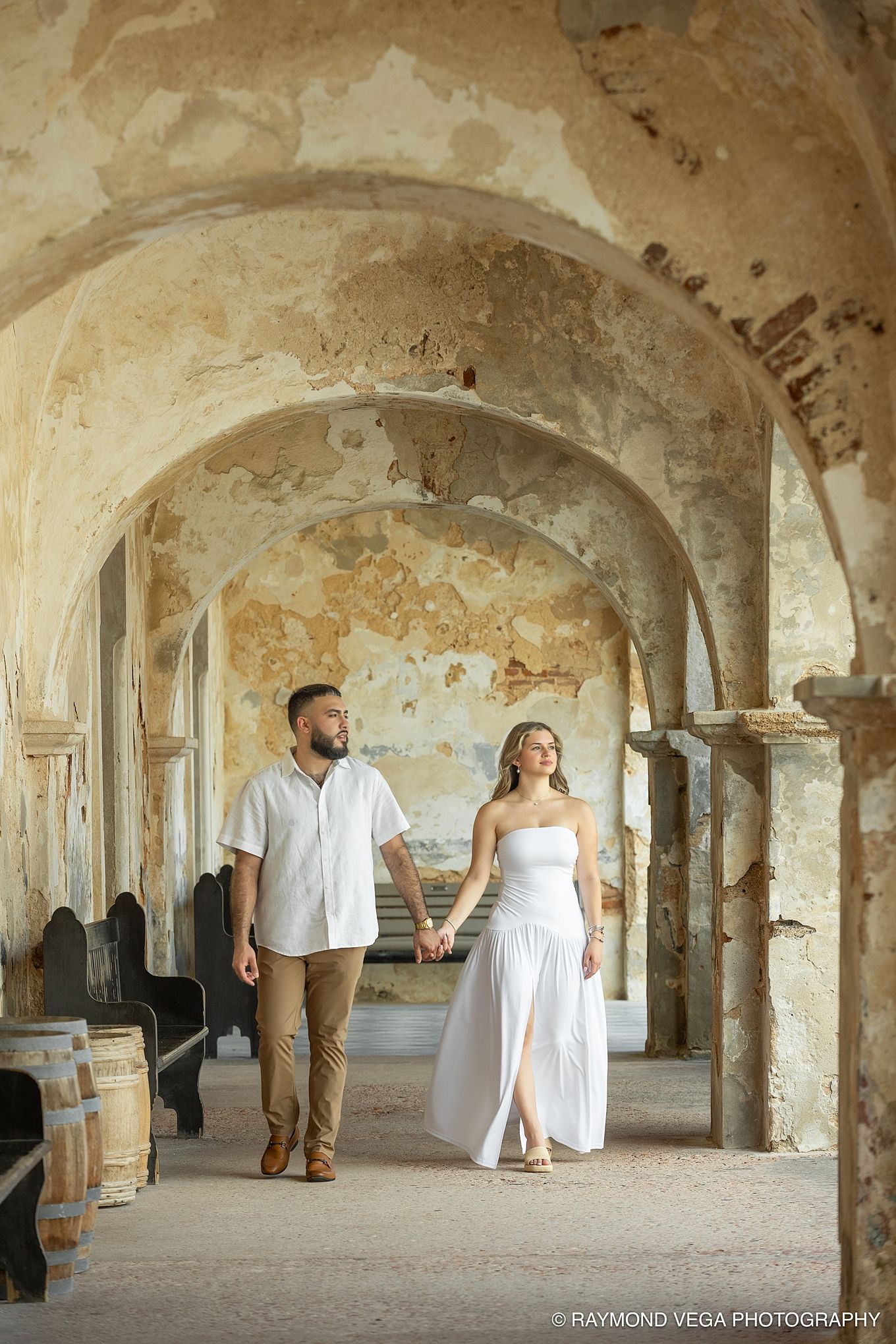 Couple holding hands walking through rustic arched stone corridor during engagement photo session.