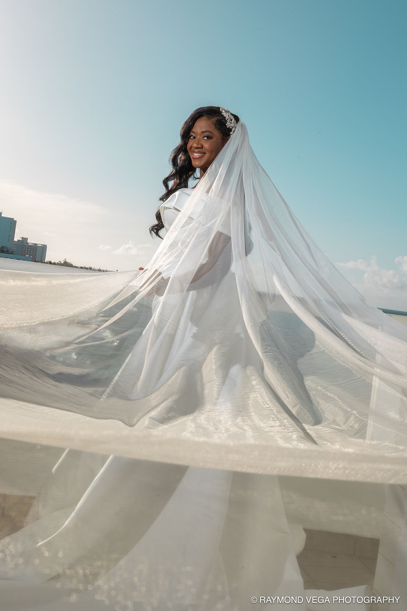 Bride in flowing white gown and veil poses gracefully on a sunny beach, with her dress billowing in the wind. Hotel Condado Vanderbilt