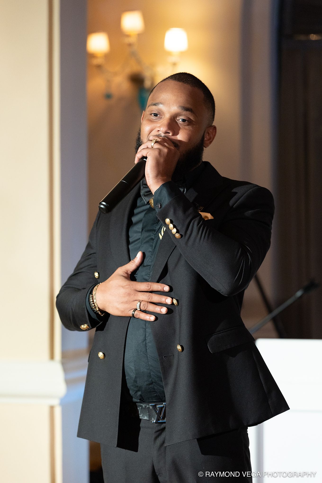 A man in a black military-style blazer holds a microphone while performing at an elegant indoor event.