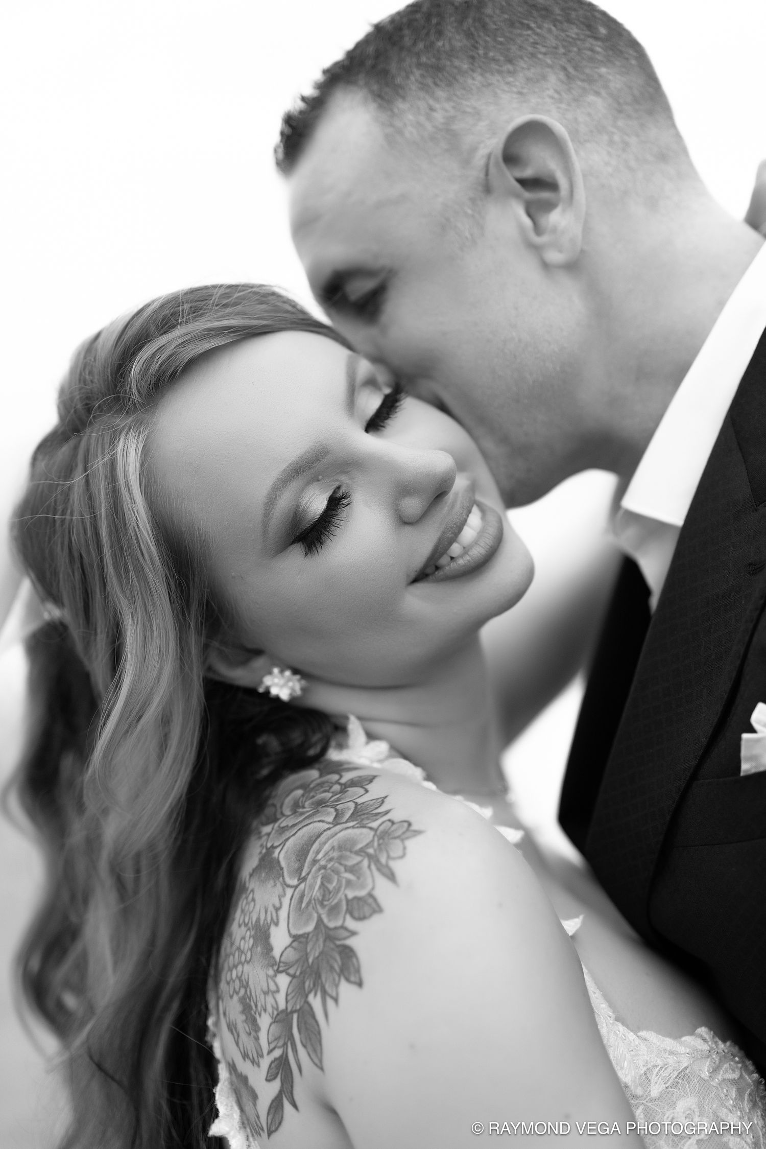 Black and white wedding photo of smiling bride with floral shoulder tattoo and groom kissing her cheek. Raymond Vega Photography & Cinema