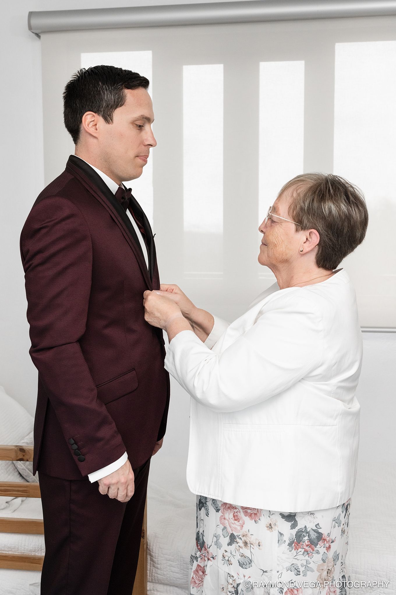 Elderly woman adjusting a young man's boutonniere on his burgundy tuxedo jacket before a formal event.