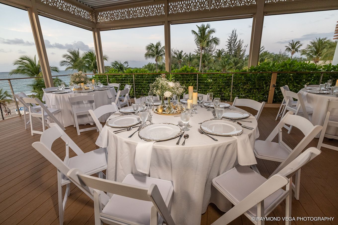 Elegant outdoor wedding reception on a tropical terrace with white round tables, chairs, and ocean views at sunset at Courtyard by Marriott