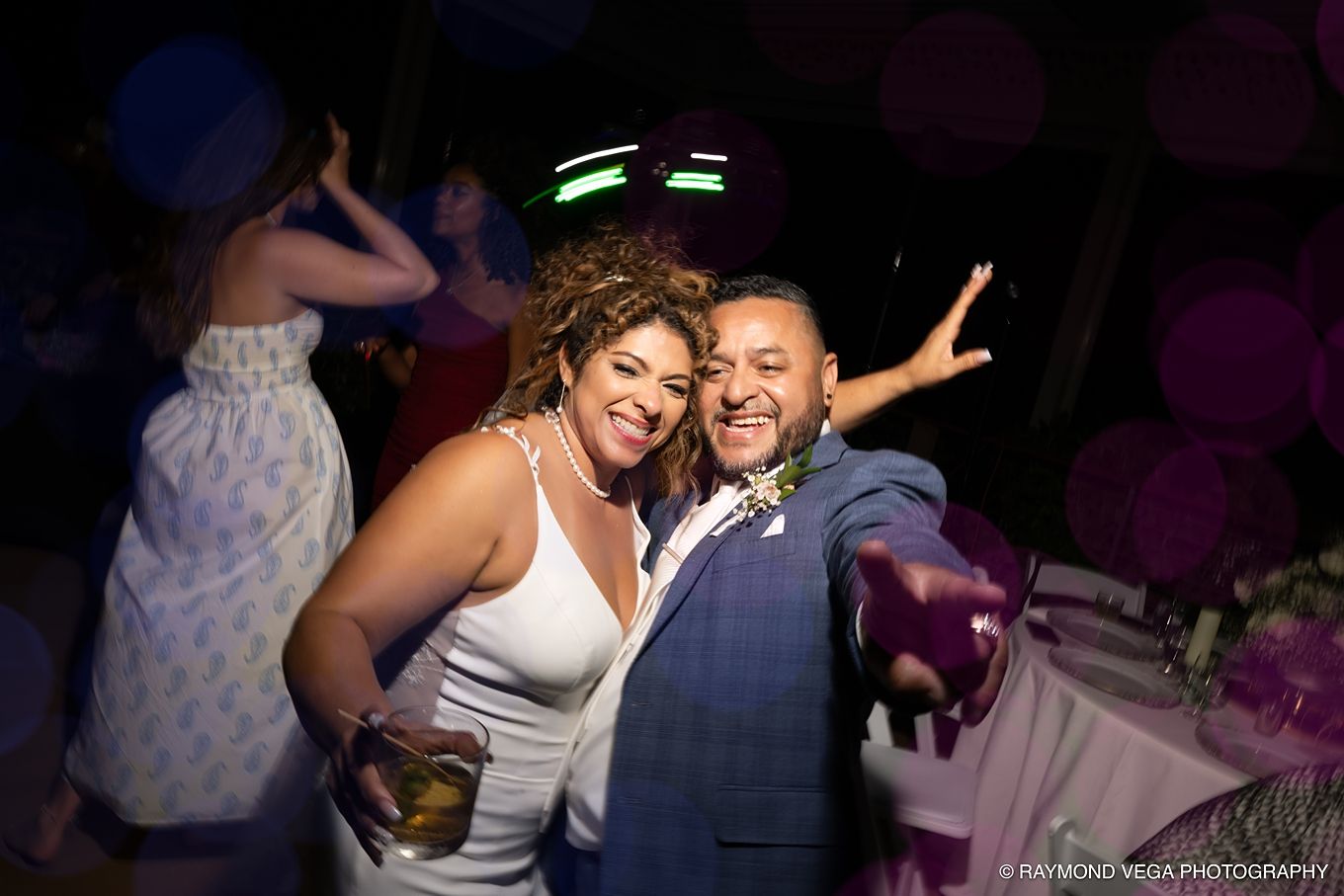 Couple smiling and posing together at a wedding reception party with purple lighting and balloons in background.