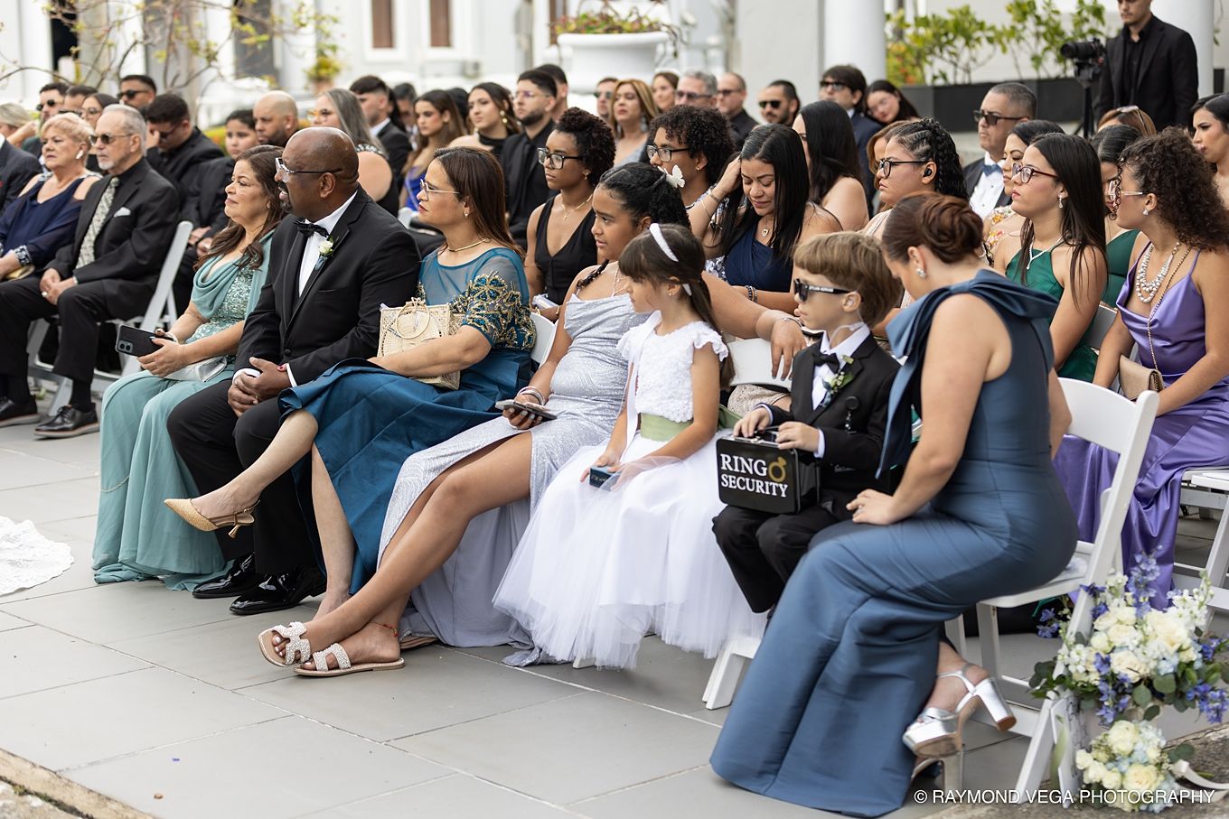 Guests seated at an outdoor wedding ceremony, including a flower girl in white dress and attendees in formal attire. Antiguo Casino