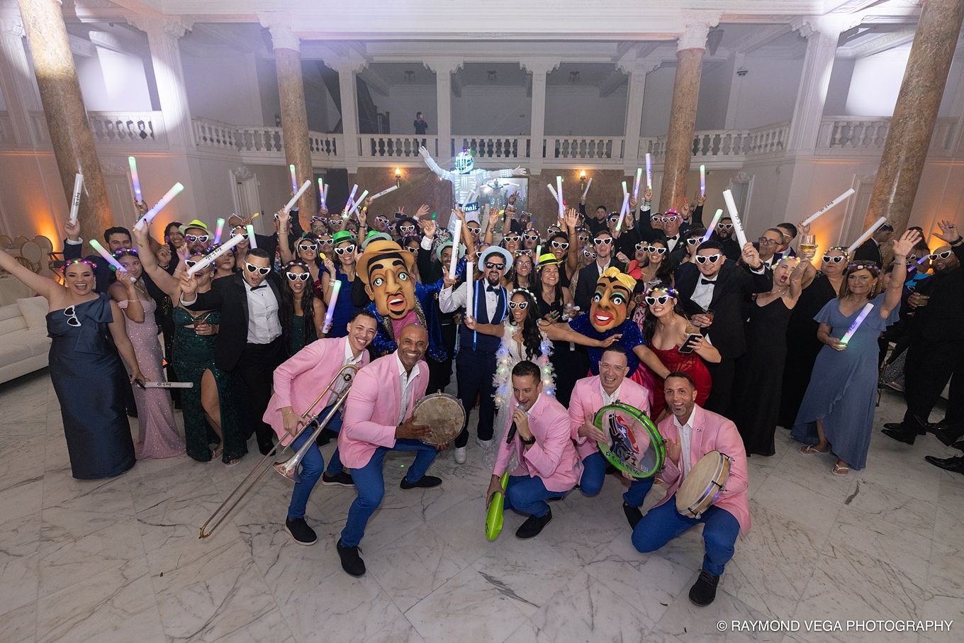 Large festive group celebrating indoors with colorful masks, glowing sticks, and formal attire in an elegant ballroom venue. Pleneros PR