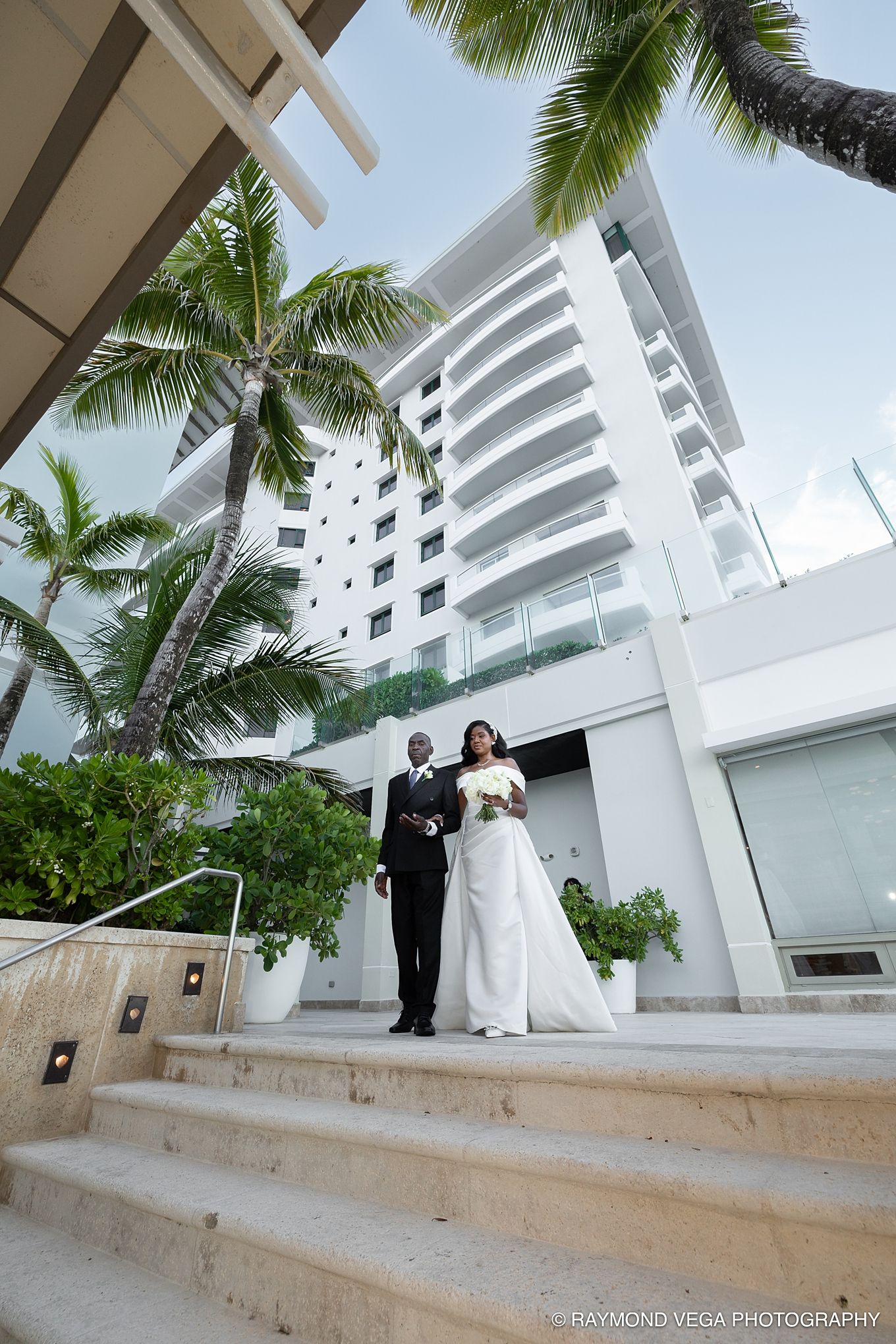 Bride and groom descend elegant stone steps at a tropical beachfront hotel, surrounded by palm trees. Vanderbilt Hotel Wedding