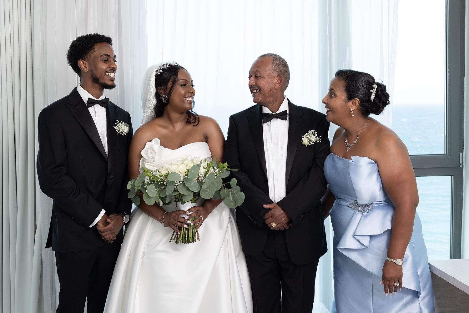 Bride in white gown holds bouquet with groom in tuxedo and family in formal wear posing indoors near window.
