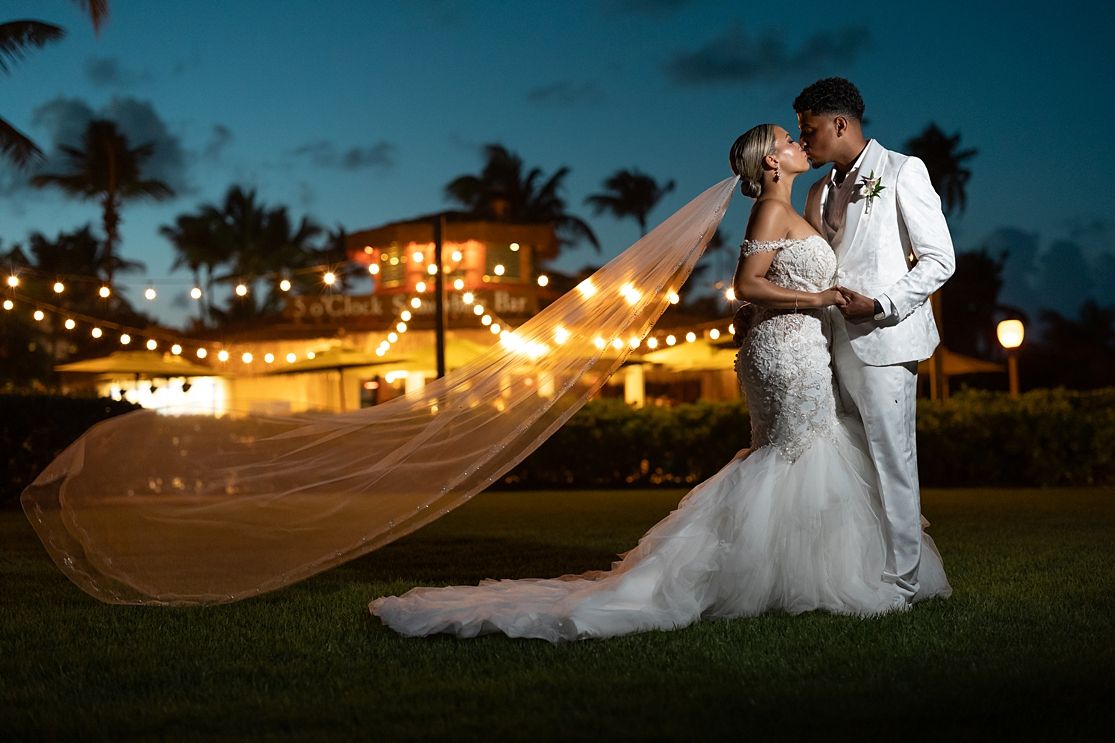 A stunning bride and groom share a romantic kiss at dusk with string lights and palm trees at a tropical wedding venue.
