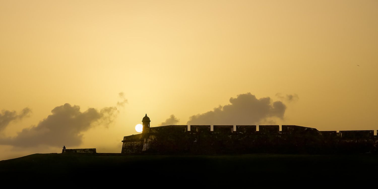 Silhouette of a historic fort against a golden sunset sky with dramatic clouds in the background.