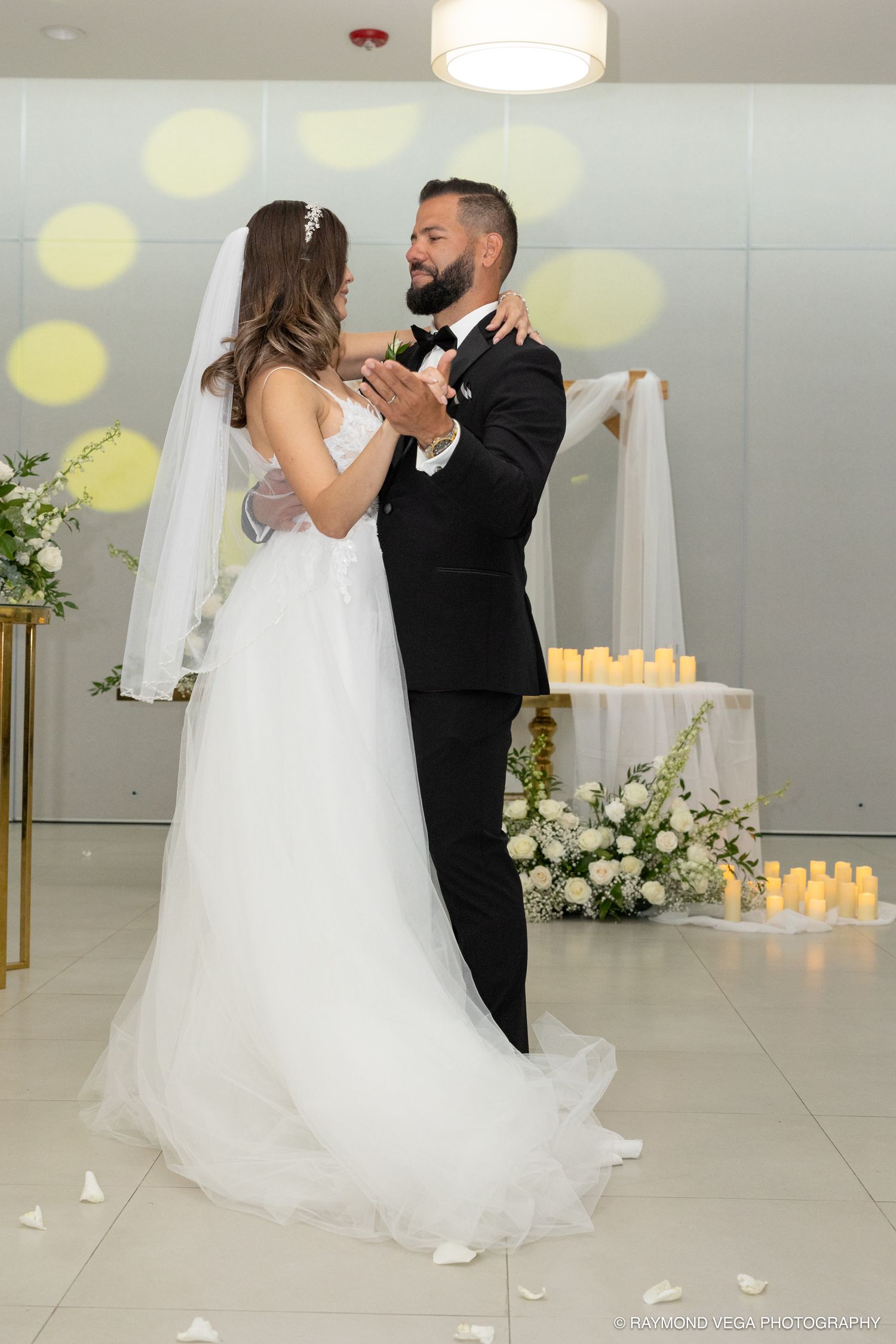 Bride and groom share their first dance, she in a white gown with veil, he in a black tuxedo, candles and florals behind.