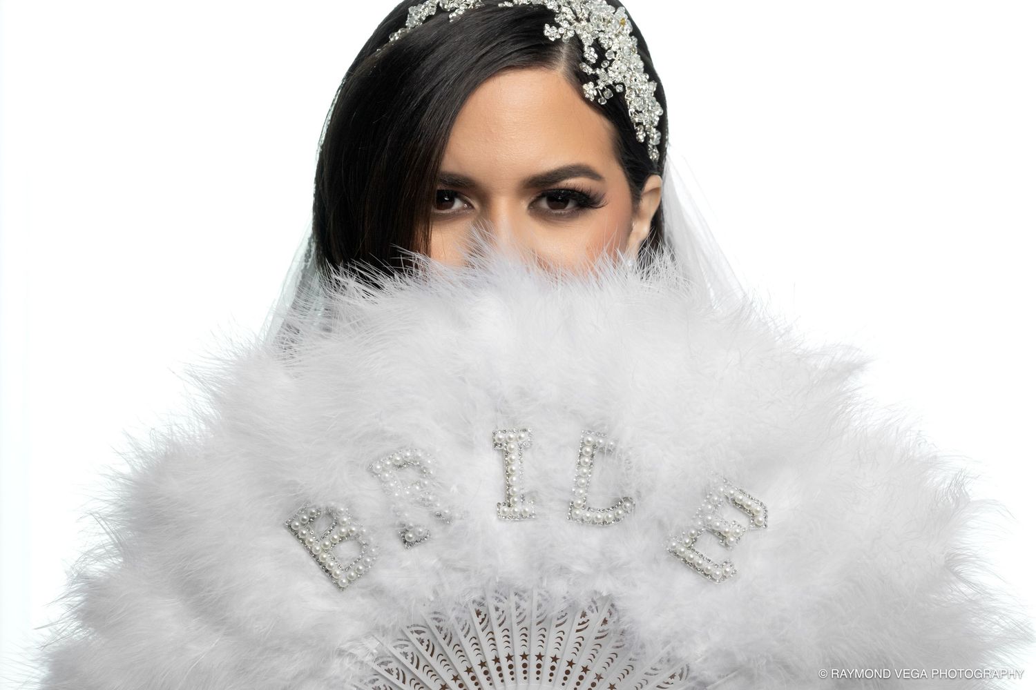 Woman holding a white feather bridal fan with rhinestone BRIDE text, wearing an ornate crystal headpiece.