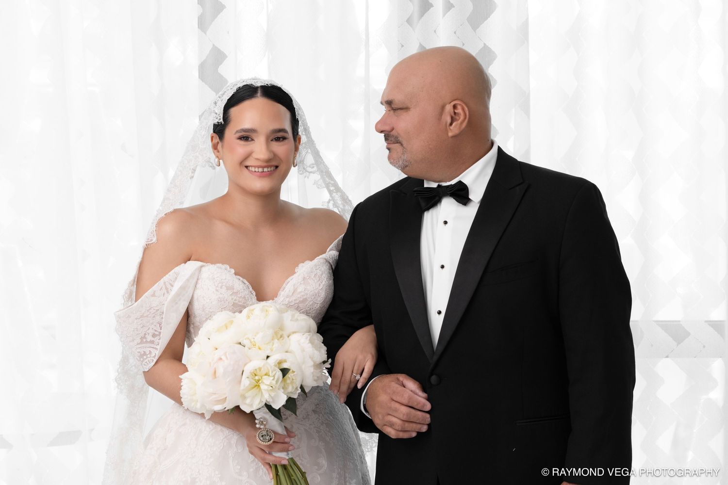 Bride in white off-shoulder gown holding bouquet stands with bald man in tuxedo at elegant wedding venue.