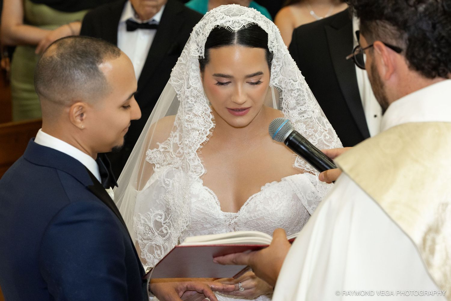 Bride in lace wedding gown and veil exchanges vows with groom in blue suit during church wedding ceremony.
