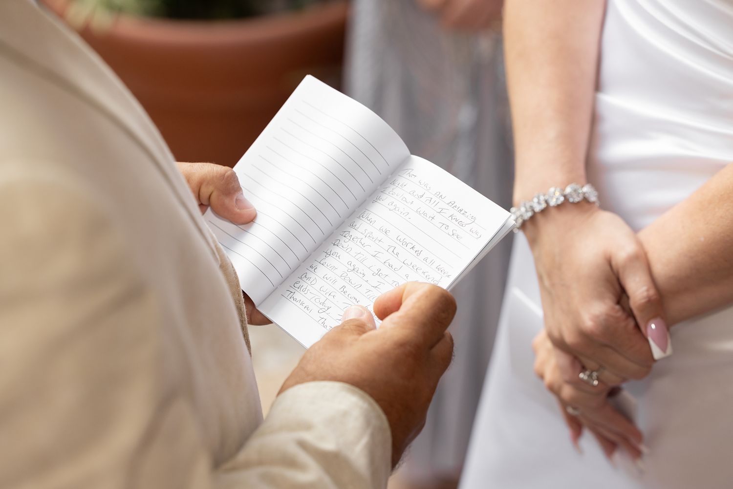 Groom reading handwritten wedding vows from a small notebook during an outdoor ceremony with bride in white dress.