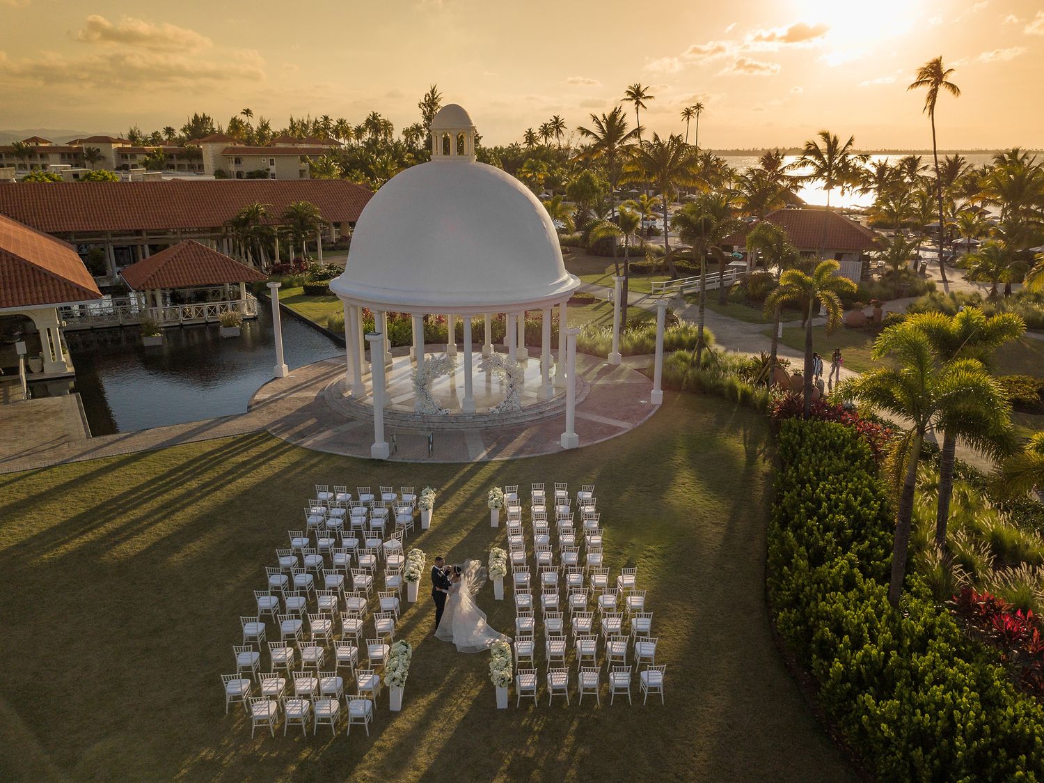 Aerial sunset view of a white gazebo wedding ceremony setup with chairs on a tropical resort lawn near the ocean.
