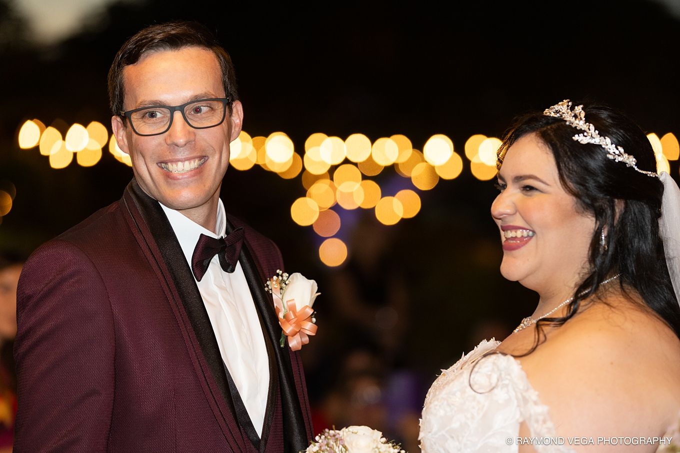 Happy bride and groom smiling at outdoor wedding reception with bokeh lights in background.
