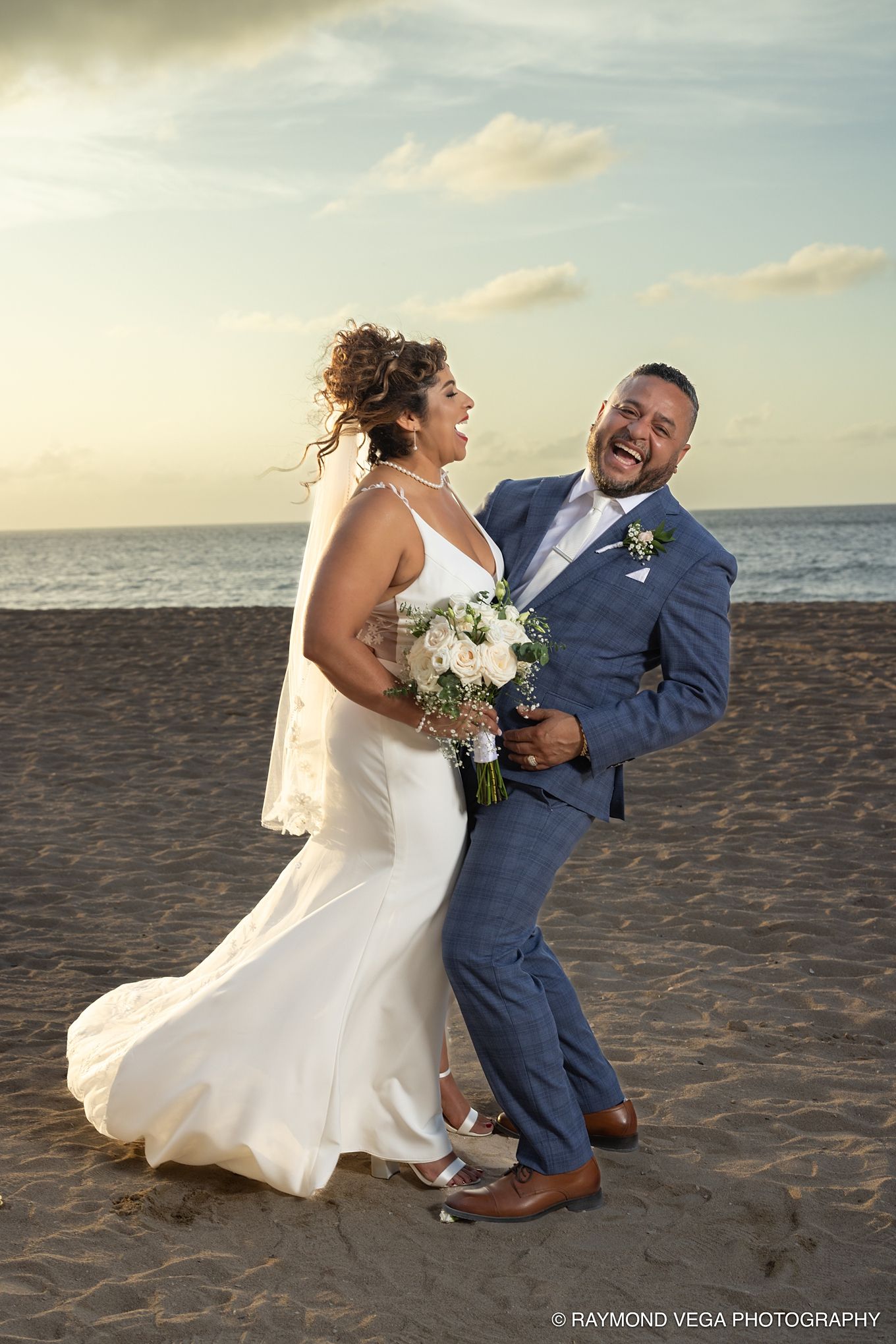 Laughing bride and groom embrace on a beach at sunset, she holds white bouquet, he wears blue suit, ocean backdrop. at Courtyard by Marriott