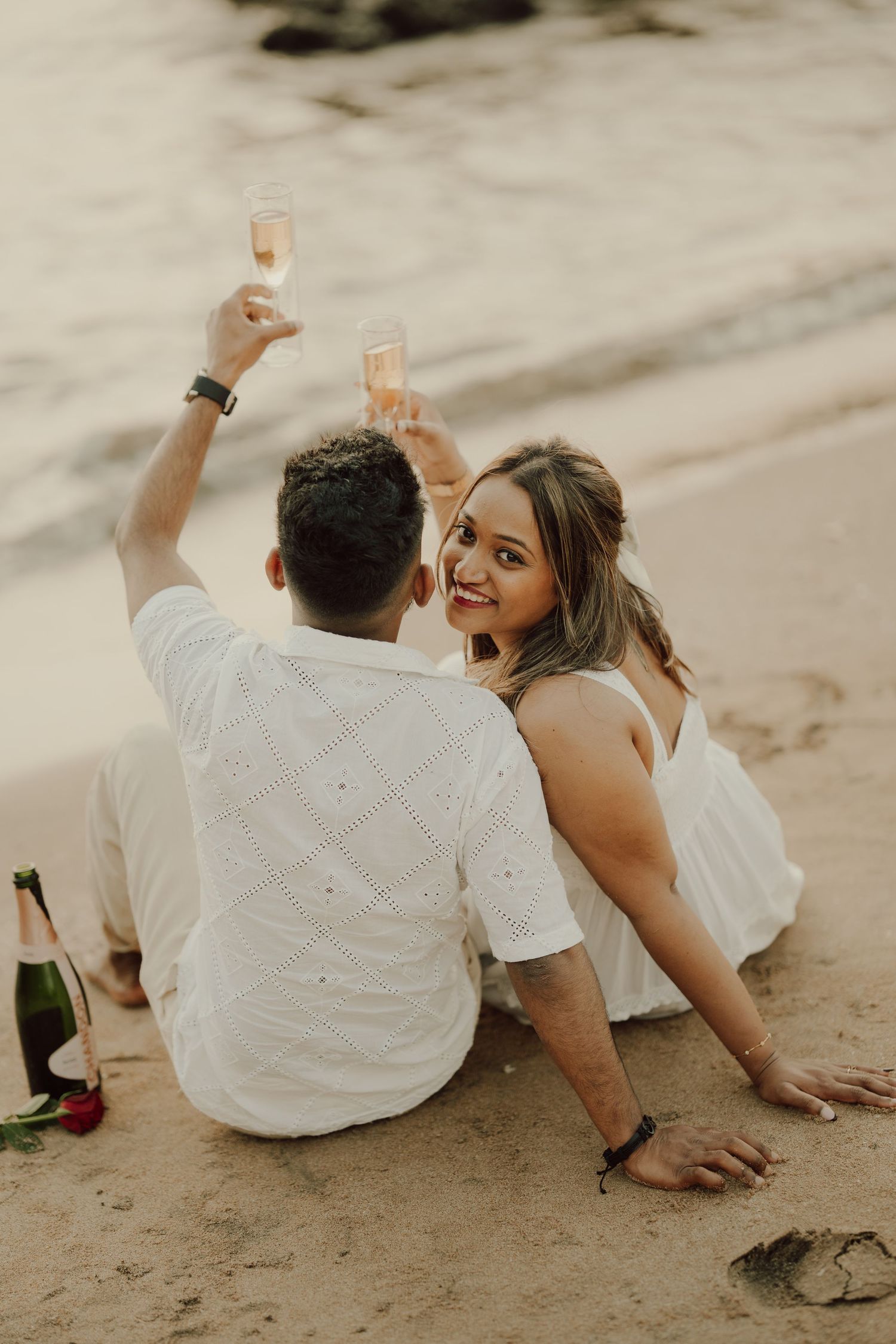 Couple in white outfits sitting on sandy beach while sharing drinks and having a romantic moment together.