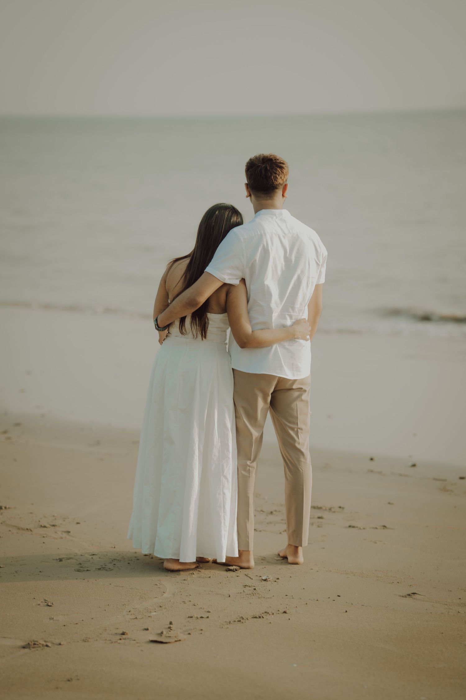 A couple in light-colored clothing embraces while standing on a misty beach at sunset.