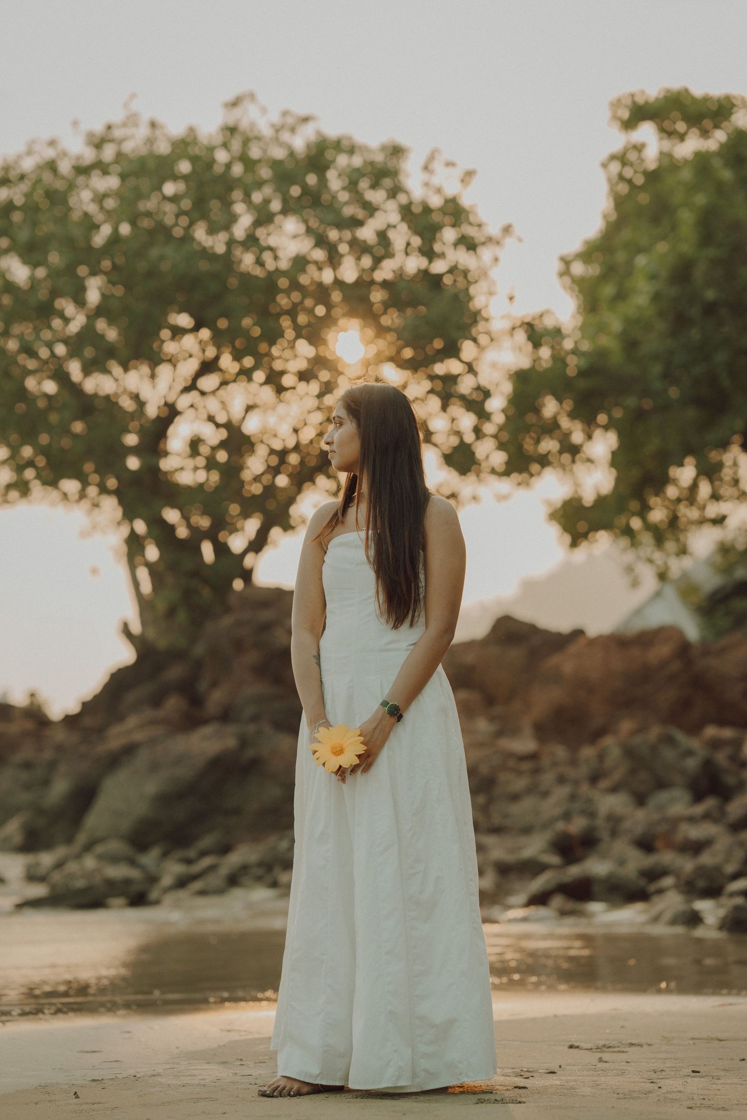 A person in a flowing white dress stands on a rocky beach at sunset with trees silhouetted in the background.