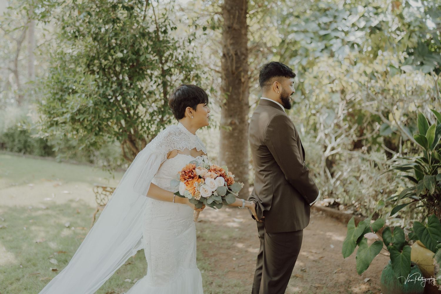 Wedding couple standing on garden path before first look reveal, with bride holding colorful bouquet of flowers.