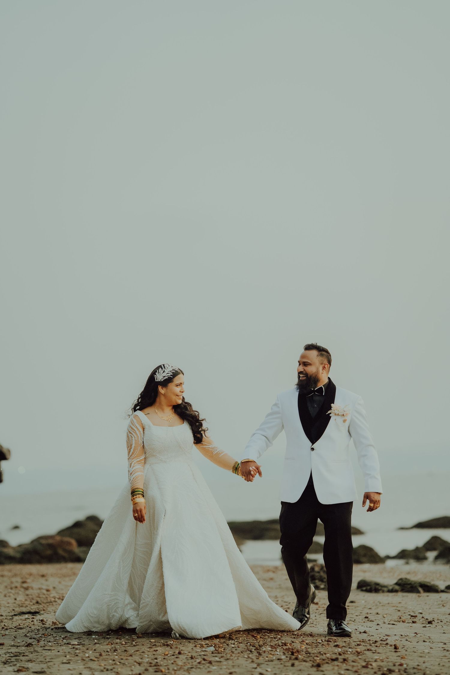 A couple holds hands and walks on a beach during their wedding photoshoot, dressed in white wedding attire.