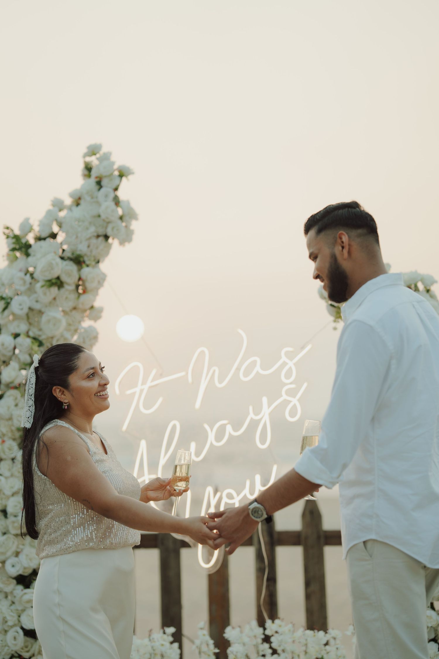 Couple holding champagne glasses at a romantic outdoor wedding ceremony with white floral arch and neon sign.
