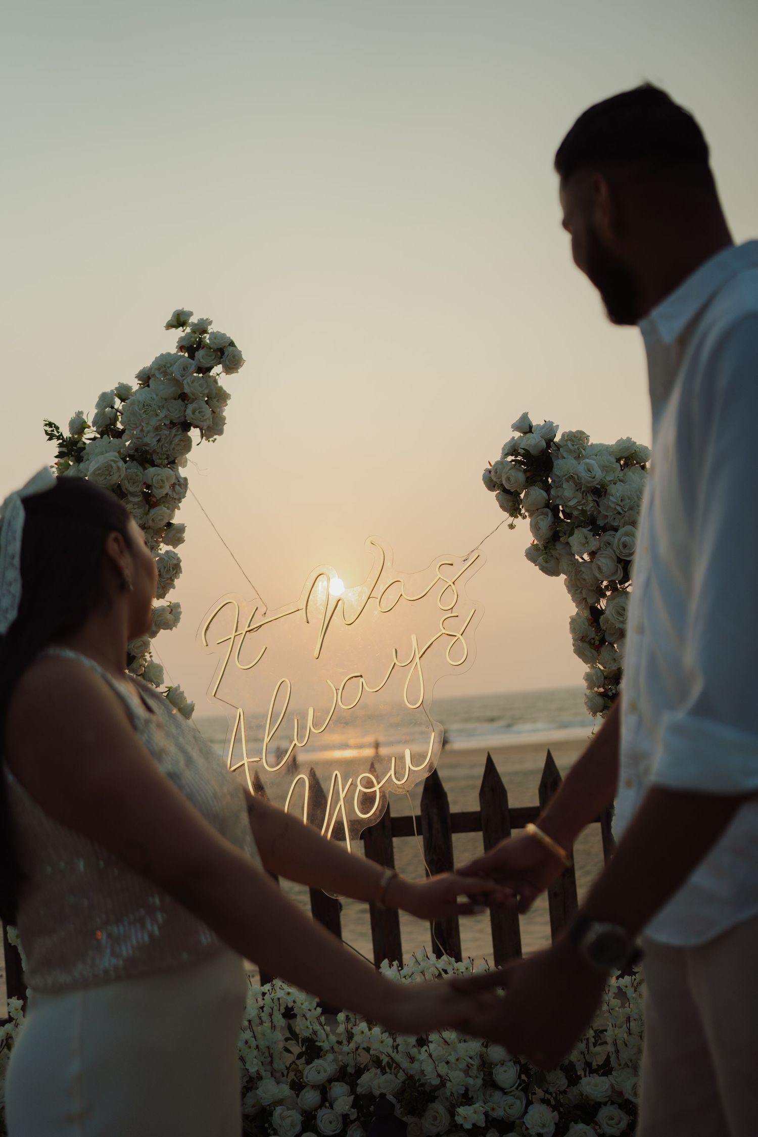 Couple holding hands at sunset beach wedding with white floral arch and neon sign reading It Was Always You.
