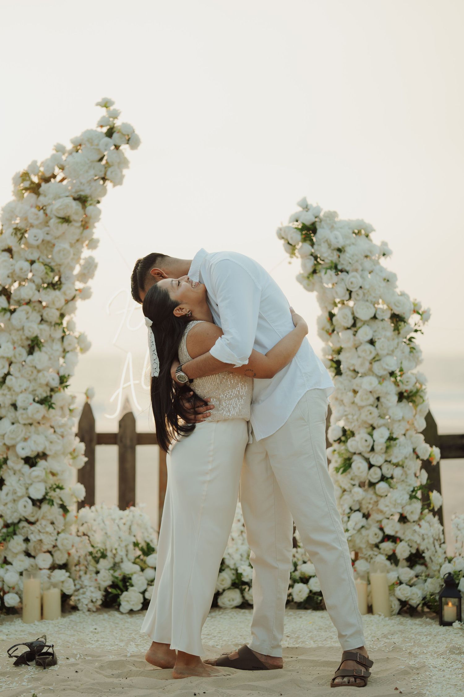 Couple sharing a romantic kiss surrounded by white floral columns on a sandy beach at sunset.